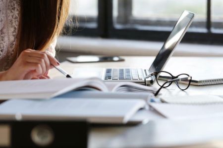 Laptop, okulary, osoba trzymająca długopis w dłoni